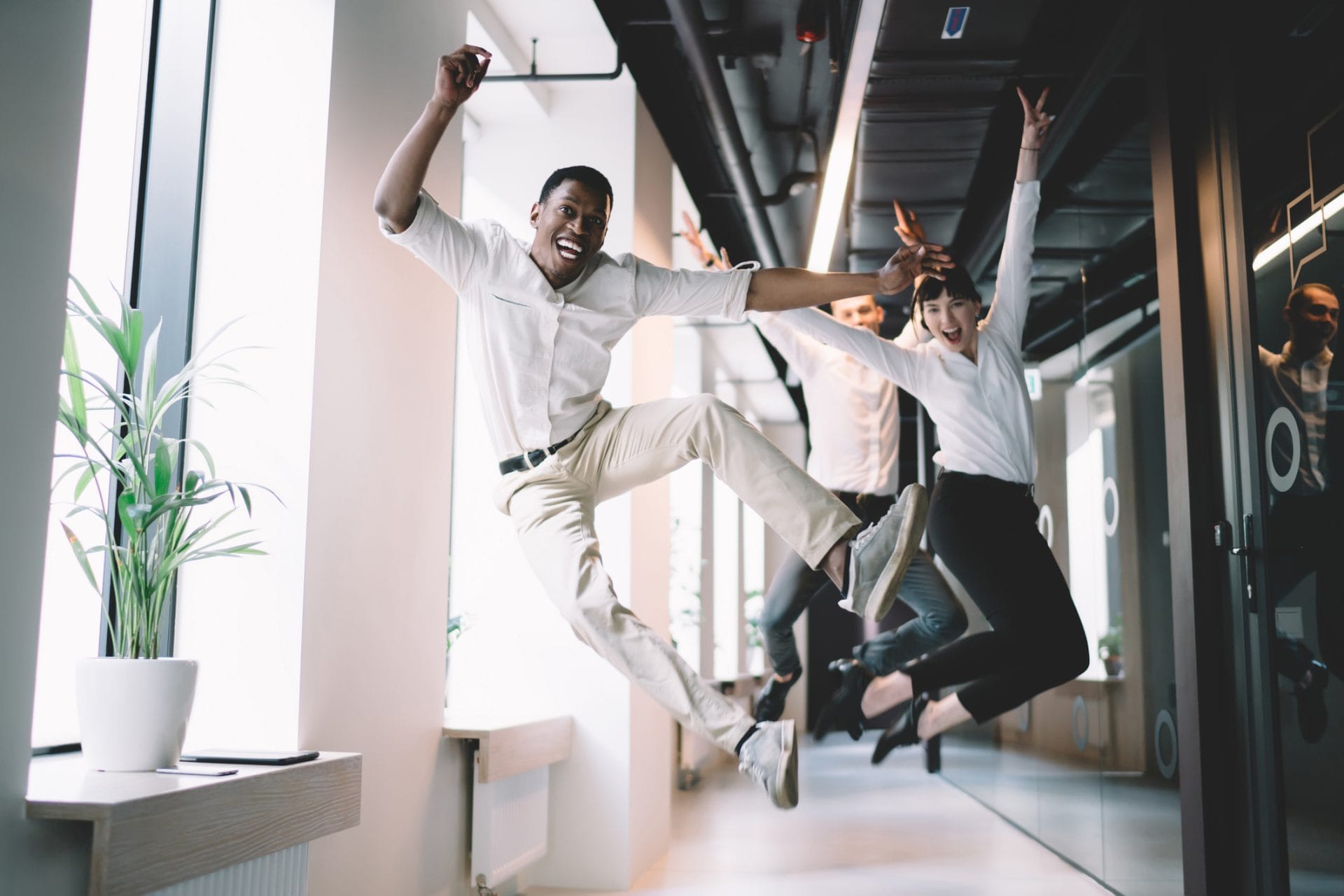 Three coworkers jumping in celebration in a bright modern office hallway